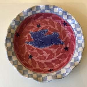 May include: A round ceramic plate with a scalloped edge featuring a blue and white checkered border. The center is painted with a red leaf pattern, dark blue stars, and a central dark blue rabbit figure in a resting position on the surface.