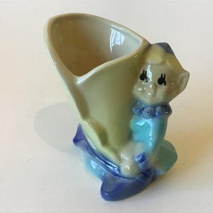 May include: A vintage ceramic figurine in the shape of a shoe, with a seated elf figure. The shoe is pale yellow, and the elf is wearing a blue outfit with a ruffled collar. The base and accents are dark blue.