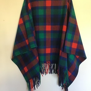 May include: A poncho featuring a vibrant plaid pattern in shades of green, blue, black, and bright red. The garment has a soft, textured appearance and is finished with long, dangling fringe along the bottom hem, hanging from a white hanger.