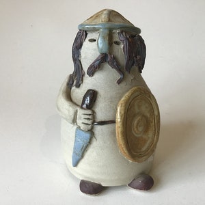 May include: A ceramic figurine of a warrior with a helmet, shield, and sword. The figurine is painted in shades of brown, blue, and white.