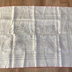 May include: Off-white linen fabric with a floral basket design. The design features a basket filled with roses and other flowers, outlined in black. The fabric has a red border and text at the top.