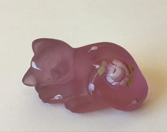 Puede incluir: Una figura de cristal esmerilado de color malva que representa a un gato durmiendo. El gato aparece en posición acurrucada con un diseño floral pintado a mano que incluye una rosa rosa claro con hojas verdes y pequeños detalles blancos.