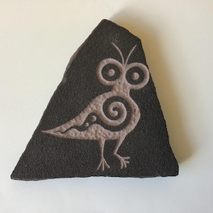 May include: A triangular dark gray stone slab featuring a light-colored engraved design of a stylized bird. The bird has large circular eyes, a spiral pattern on its body, and thin legs. The surface of the stone has a rough, textured appearance.