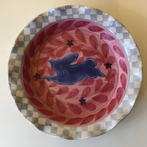 May include: A round ceramic bowl with a scalloped edge featuring a blue and white checkered pattern. The interior displays a central blue rabbit graphic surrounded by red leaf-like brushstrokes and small dark blue stars on a light red background.