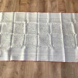 May include: Rectangular off-white fabric with a red border, featuring a detailed floral design in gray. The fabric is folded and has text at the bottom. The design includes leaves and stems.