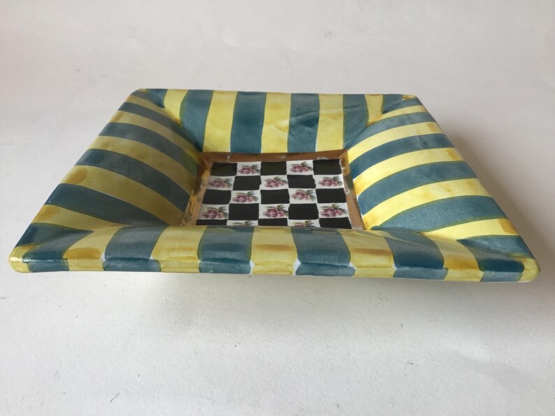 May include: Square decorative dish with a yellow and teal striped border. The center of the dish features a checkerboard pattern with pink floral accents. The dish has a slightly raised edge.