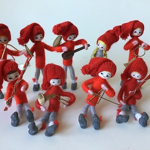 May include: A collection of handmade Christmas elf ornaments. Each elf is dressed in a red knitted outfit with a matching hat. Some elves are playing musical instruments, including a guitar, trumpet, and violin. The elves have gray legs and black shoes.