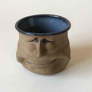 May include: A small, round, tan-colored ceramic cup with a textured, matte exterior surface. The cup features a sculpted human-like face with a wide, smiling mouth and closed eyes. The interior of the cup is glazed in a smooth, deep blue finish.