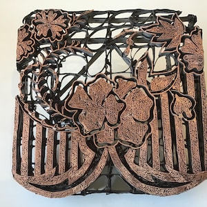 May include: A decorative, square-shaped object with a floral design. The object features a black, openwork frame with copper-colored floral and leaf accents. The design includes a large flower in the center and vertical stripes.