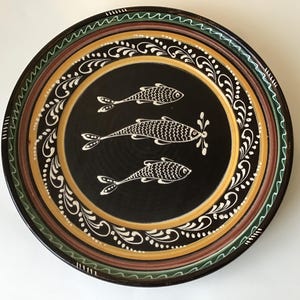 May include: A round, decorative ceramic plate with a black center featuring three white fish. The plate has a white, yellow, and brown border with a decorative white floral pattern. The outer rim is green and brown.