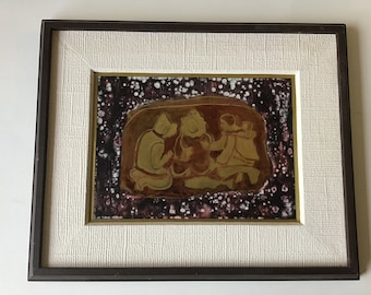 May include: A framed artwork featuring a relief sculpture of three figures in a gold color against a dark background with white speckles. The figures are depicted in a seated position, with one figure holding a small object.
