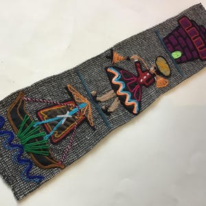 May include: A rectangular grey woven fabric strip featuring colorful embroidered scenes. The design includes a brown boat on blue wavy water, a figure in a black dress with orange and blue trim, and a purple brick structure at the top end.