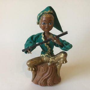 May include: A vintage figurine of a seated figure playing a flute. The figure wears a green velvet outfit with gold trim and a matching hat. The base is brown and gold, with a textured appearance.