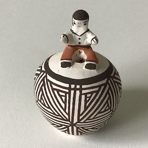 May include: A small, round, white ceramic pot with a black and white geometric design. A small, brown and white figurine of a person is sitting on top of the pot.