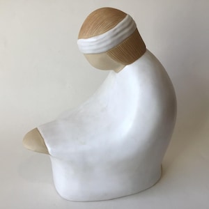 May include: A ceramic sculpture of a figure in a white robe, head bowed. The head has light brown hair and a white headband. The figure is in a seated position, with the knees bent.