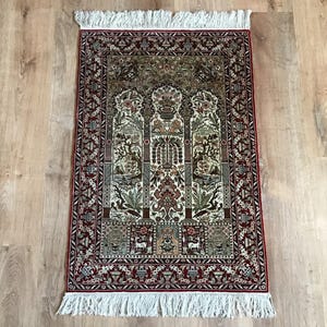 May include: A rectangular, ornate rug with a central design featuring three arched panels with floral and geometric patterns. The rug has a red border and white fringe. The color palette includes red, green, brown, and cream.