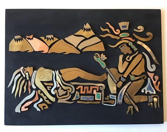 May include: A decorative wall art piece featuring an Aztec-inspired design. The artwork is composed of various metal pieces in shades of gold, copper, and silver, set against a black background. The scene depicts figures, mountains, and stylized elements.