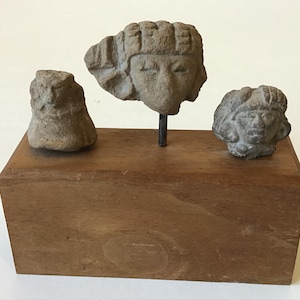 May include: Three weathered, gray stone sculptures of human heads and a small form are displayed on a wooden block. The central head is mounted on a metal rod. The sculptures appear to be ancient artifacts.