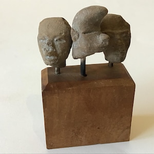 May include: A small, brown wooden base supports three weathered, stone-like sculptures of human heads. The heads are attached to the base with metal rods. The sculpture has a rustic, antique appearance.