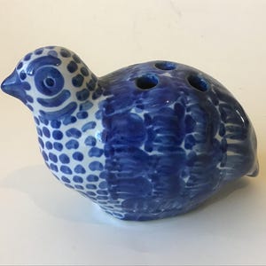 May include: A ceramic bird figurine in shades of blue and white. The bird has a speckled head and a body with a textured, painted design. Three holes are visible on the top, suggesting a decorative or functional purpose.