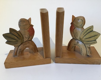 May include: A pair of wooden bookends featuring carved birds with green, brown, and red accents. The birds are perched on a branch and have their beaks open as if singing.