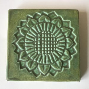 May include: Square, green ceramic tile with a raised sunflower design. The sunflower is centered on the tile and has detailed petals and a textured center. The tile has a matte finish and is likely for decorative use.