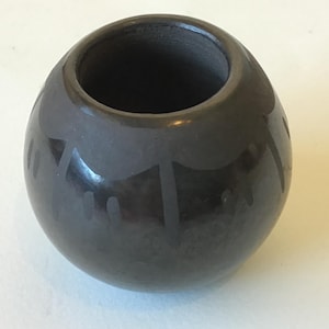 May include: A dark gray, round ceramic vase with a wide opening. The vase has a glossy finish and is decorated with a simple, repeating pattern around the body. The design appears to be etched or painted onto the surface.
