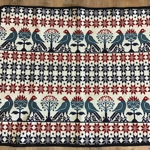 May include: A square woven blanket featuring a repeating pattern of birds and stylized trees in dark blue and red on an off-white background. The design includes horizontal bands of geometric star and diamond shapes with a solid black border.