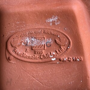 May include: Close-up of a terracotta-colored ceramic object with an oval emblem. The emblem reads "Victoria and Richard" above "Mackenzie-Childs, Ltd." and "1995." The object has a textured surface and appears to be a base or underside of a decorative piece.