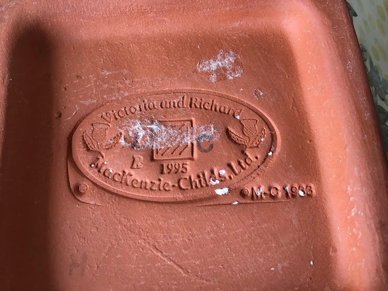 May include: Close-up of a terracotta-colored ceramic object with an oval emblem. The emblem reads "Victoria and Richard" above "Mackenzie-Childs, Ltd." and "1995." The object has a textured surface and appears to be a base or underside of a decorative piece.