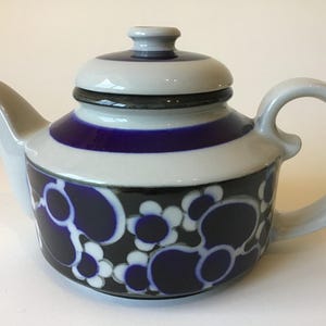 May include: A ceramic teapot with a light gray base color, featuring a dark blue band around the upper body and a lid. The lower section is decorated with a repeating pattern of dark blue and white circular flower shapes on a black background.