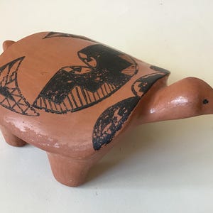 May include: A terracotta-colored ceramic turtle figurine with black geometric designs on its shell. The turtle has a smooth, rounded body, a long neck, and small legs. The figurine is a decorative item.
