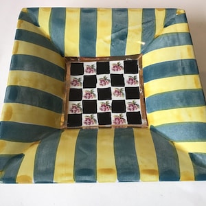 May include: Square decorative ceramic tray with a striped pattern in yellow and teal. The center features a checkerboard design with pink floral accents. The tray has a slightly curved shape and a glossy finish.