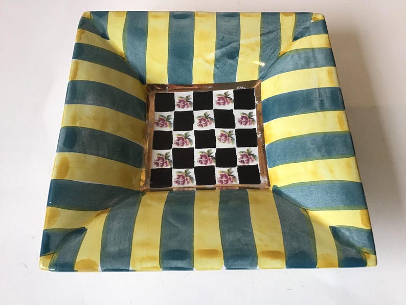 May include: Square decorative ceramic tray with a striped pattern in yellow and teal. The center features a checkerboard design with pink floral accents. The tray has a slightly curved shape and a glossy finish.