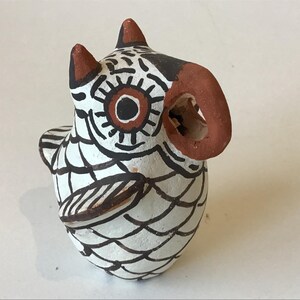 May include: A hand-painted ceramic owl figurine. The owl is primarily white with black and brown details. It features a large, circular eye with a brown center and a curved, reddish-brown beak. The owl's body is decorated with a scale-like pattern.