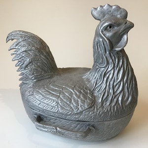 May include: A silver metal rooster-shaped serving dish with a hinged lid. The rooster is detailed with feathers and has a textured surface.