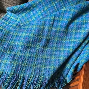 May include: A woven blanket in shades of blue and green with a textured pattern. The blanket has a fringe along the bottom edge. The blanket is draped over a chair.