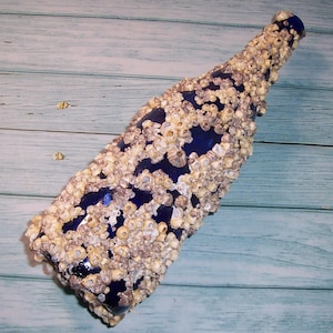 May include: A blue glass bottle encrusted with barnacles. The bottle has a long neck and a rounded body, with the barnacles covering most of its surface. The barnacles are beige and white, creating a textured appearance. The bottle is displayed on a light blue wooden surface.