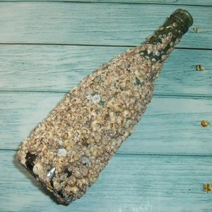 May include: A clear glass bottle encrusted with barnacles. The bottle is green at the top and covered in beige and tan barnacles. The bottle is lying on a light blue wooden surface.