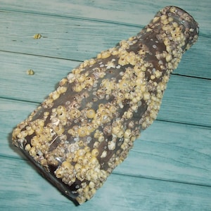 May include: A clear glass bottle covered in clusters of small, beige barnacles. The bottle is angled, showcasing its shape and the texture of the marine life attached to it. The background is a light blue wooden surface.