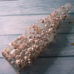 May include: A clear glass bottle decorated with a dense covering of small, light brown and white barnacle-like shells. The shells are clustered across the entire exterior surface of the bottle, creating a textured, organic appearance on the glass.