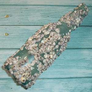 May include: A sea-worn, light green glass bottle encrusted with barnacles. The bottle's surface is covered in clusters of small, off-white and gray barnacles, creating a textured appearance. The bottle is positioned diagonally against a light blue wooden background.