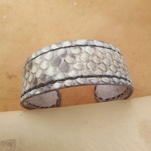 May include: A wide cuff bracelet made of light grey and white snakeskin. The bracelet has a textured pattern and is trimmed with black stitching. The open-ended design allows for easy wear. The bracelet is approximately 5 cm wide.