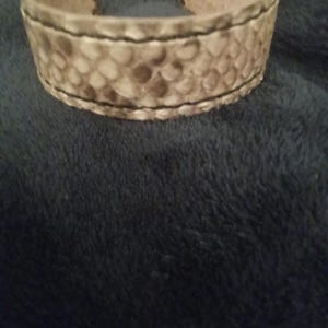 May include: A wide, open cuff bracelet with a textured, snake-like pattern in shades of tan and brown. The edges are stitched with black thread. The bracelet is made of a thick material.