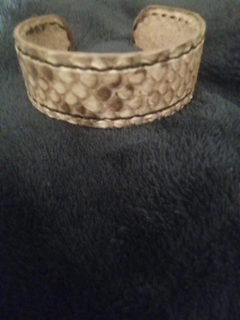 May include: A wide, open cuff bracelet with a textured, snake-like pattern in shades of tan and brown. The edges are stitched with black thread. The bracelet is made of a thick material.