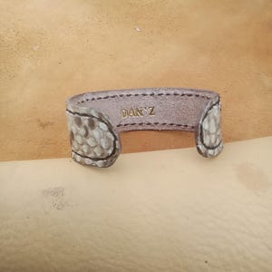 May include: A handcrafted leather cuff bracelet with a snakeskin pattern on the ends. The interior is a light brown colour, with the name "DAN'Z" embossed in gold lettering. The bracelet is open-ended.