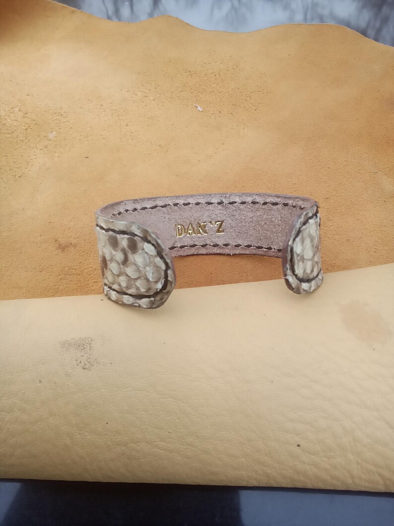 May include: A handcrafted leather cuff bracelet with a snakeskin pattern on the ends. The interior is a light brown colour, with the name "DAN'Z" embossed in gold lettering. The bracelet is open-ended.