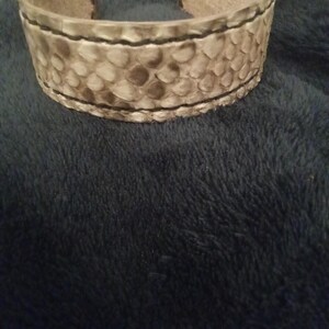 May include: A wide cuff bracelet featuring a snakeskin design. The bracelet is predominantly light grey with darker grey and brown details, and black stitching. The cuff is made from a textured material, and is open at the ends.