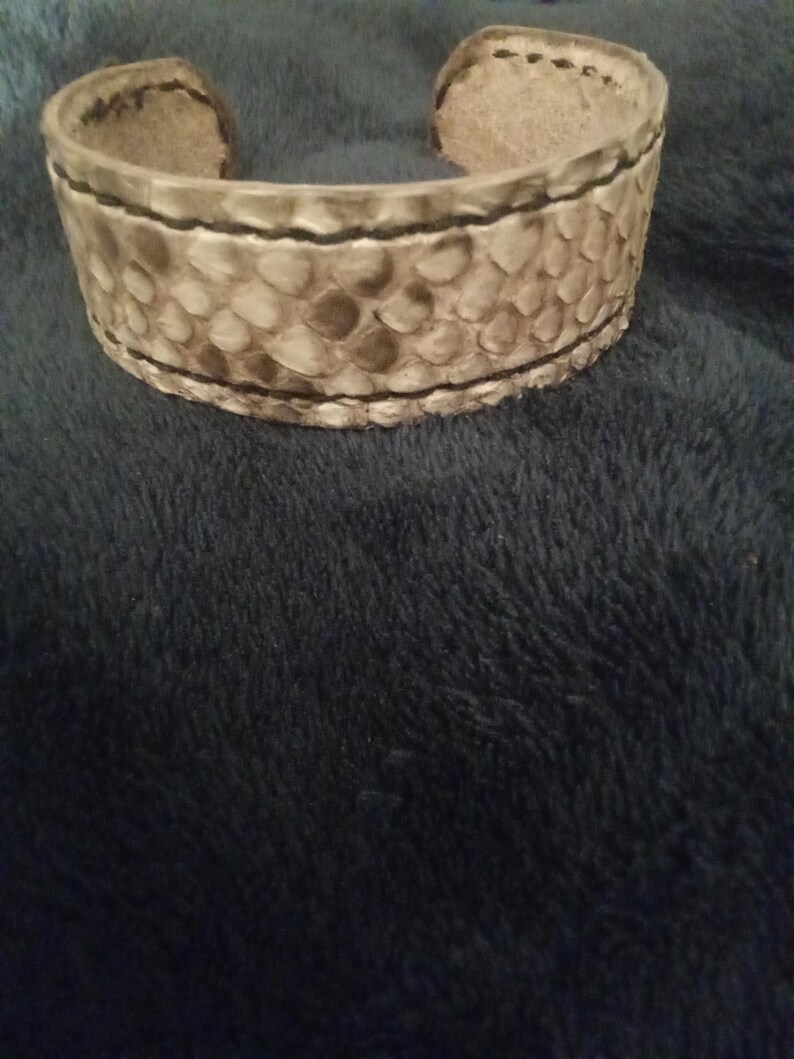 May include: A wide cuff bracelet featuring a snakeskin design. The bracelet is predominantly light grey with darker grey and brown details, and black stitching. The cuff is made from a textured material, and is open at the ends.