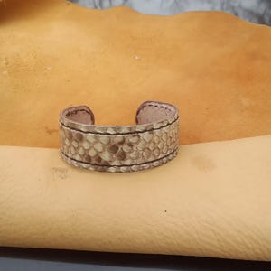 May include: A cuff bracelet made from textured, light-coloured leather with a reptile-like pattern. The bracelet has a rectangular shape with rounded edges and is accented with dark stitching. The bracelet is displayed on a tan leather surface.